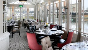 A restaurant with a high ceiling and large windows overlooking a canal. Tables are set with white tablecloths and red chairs. - Macdonald Compleat Angler (Marlow)