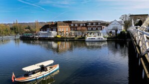 A canal with a boat docked near a bridge, a row of buildings, and a white boat in the background. - Macdonald Compleat Angler (Marlow)