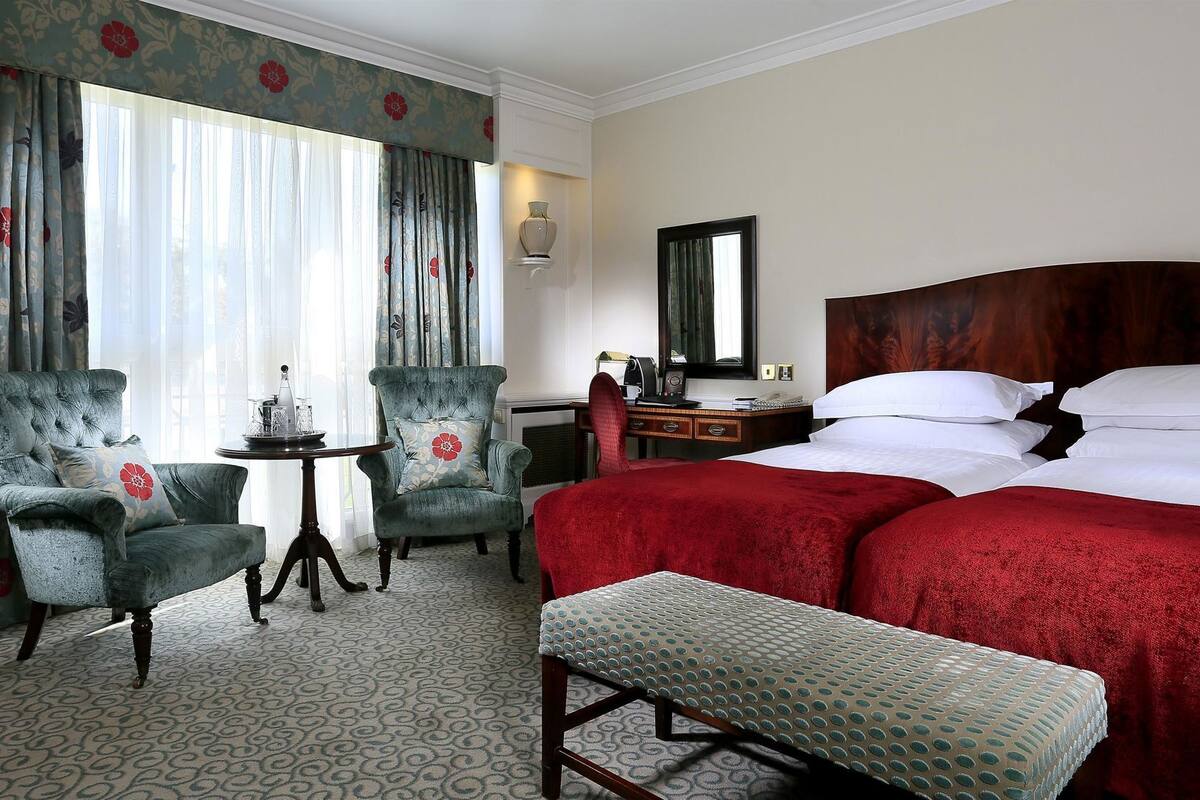 A hotel room with a large bed, two armchairs, a small table, a desk, and a bench.