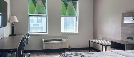 A hotel room with a bed, a desk, a chair, a TV, two windows with blinds, and a wall-mounted air conditioning unit.