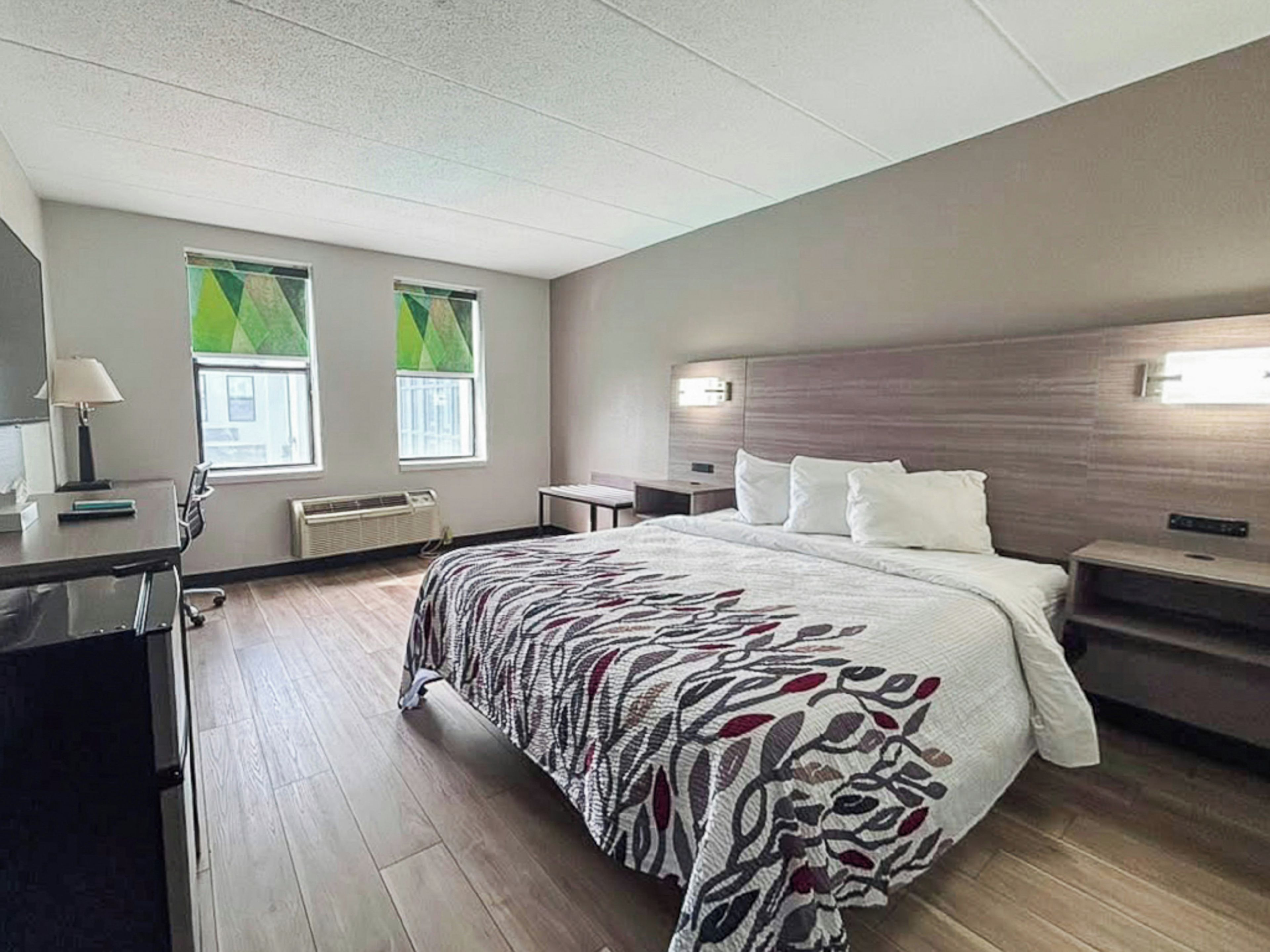Una habitación de hotel con una cama, mesitas de noche, un escritorio y una silla. Hay dos ventanas con cortinas verdes.