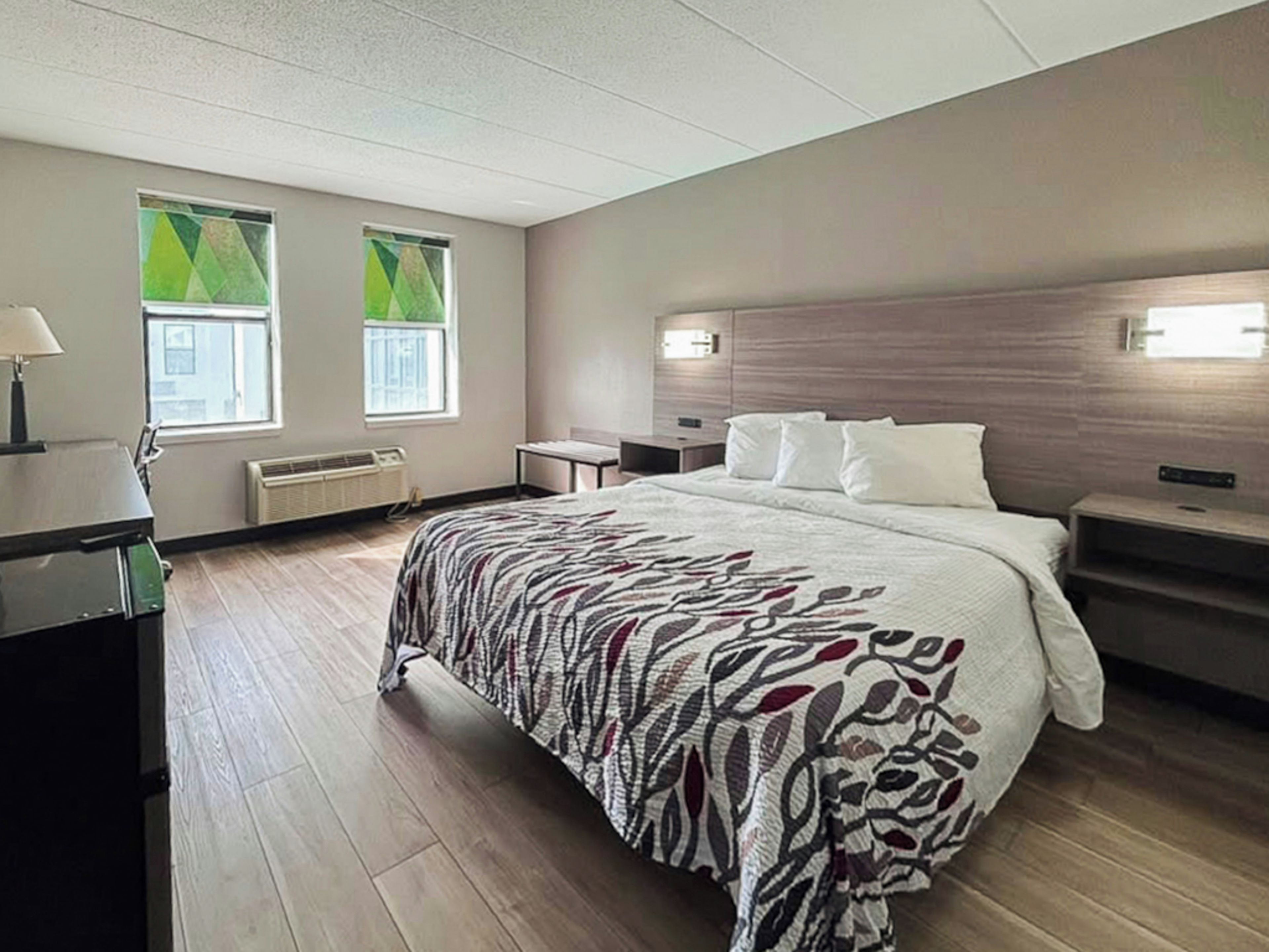 A hotel room with a large bed, bedside tables, a desk, and two windows with green curtains.