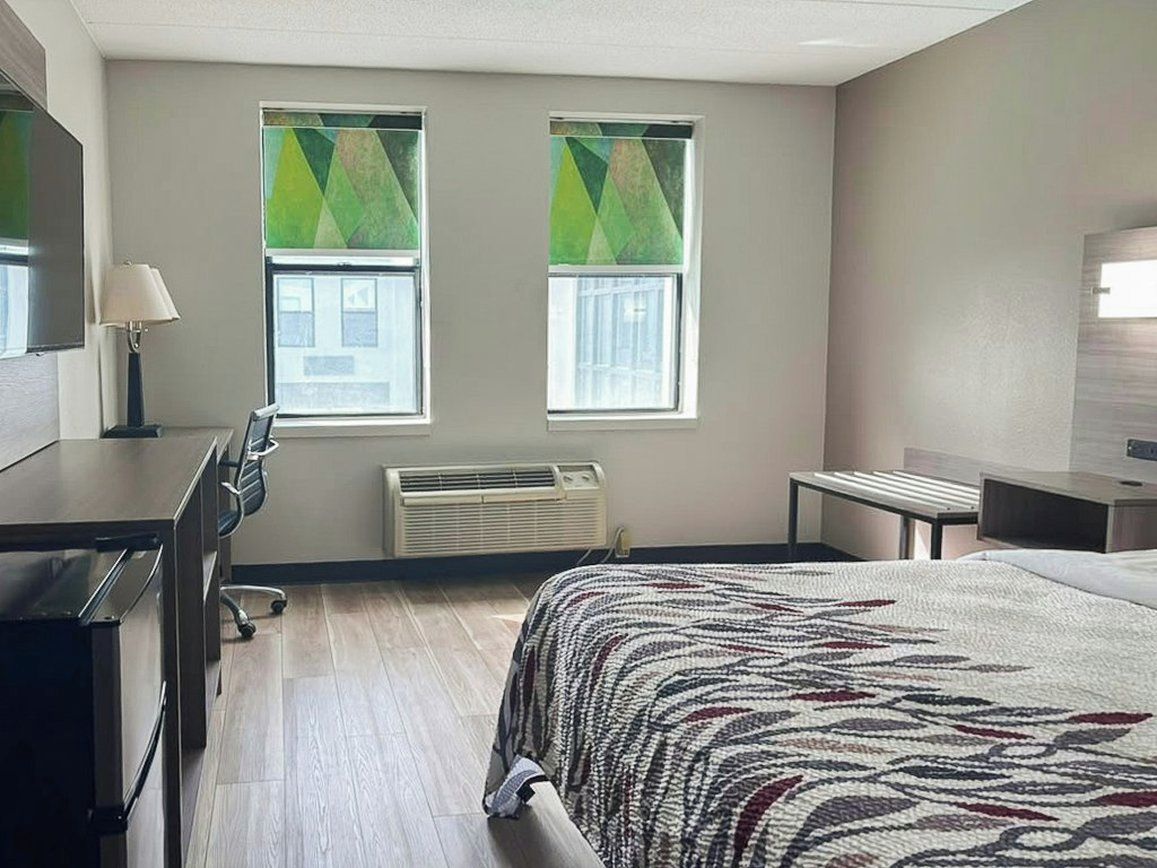 A hotel room with a bed, a desk, a chair, two windows with curtains, a heater, and a television.