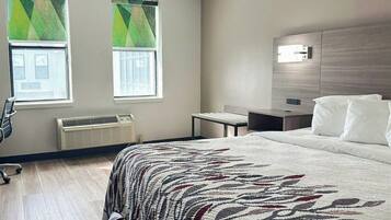 A hotel room with a bed, a desk, two windows with blinds, a heater, and a chair.
