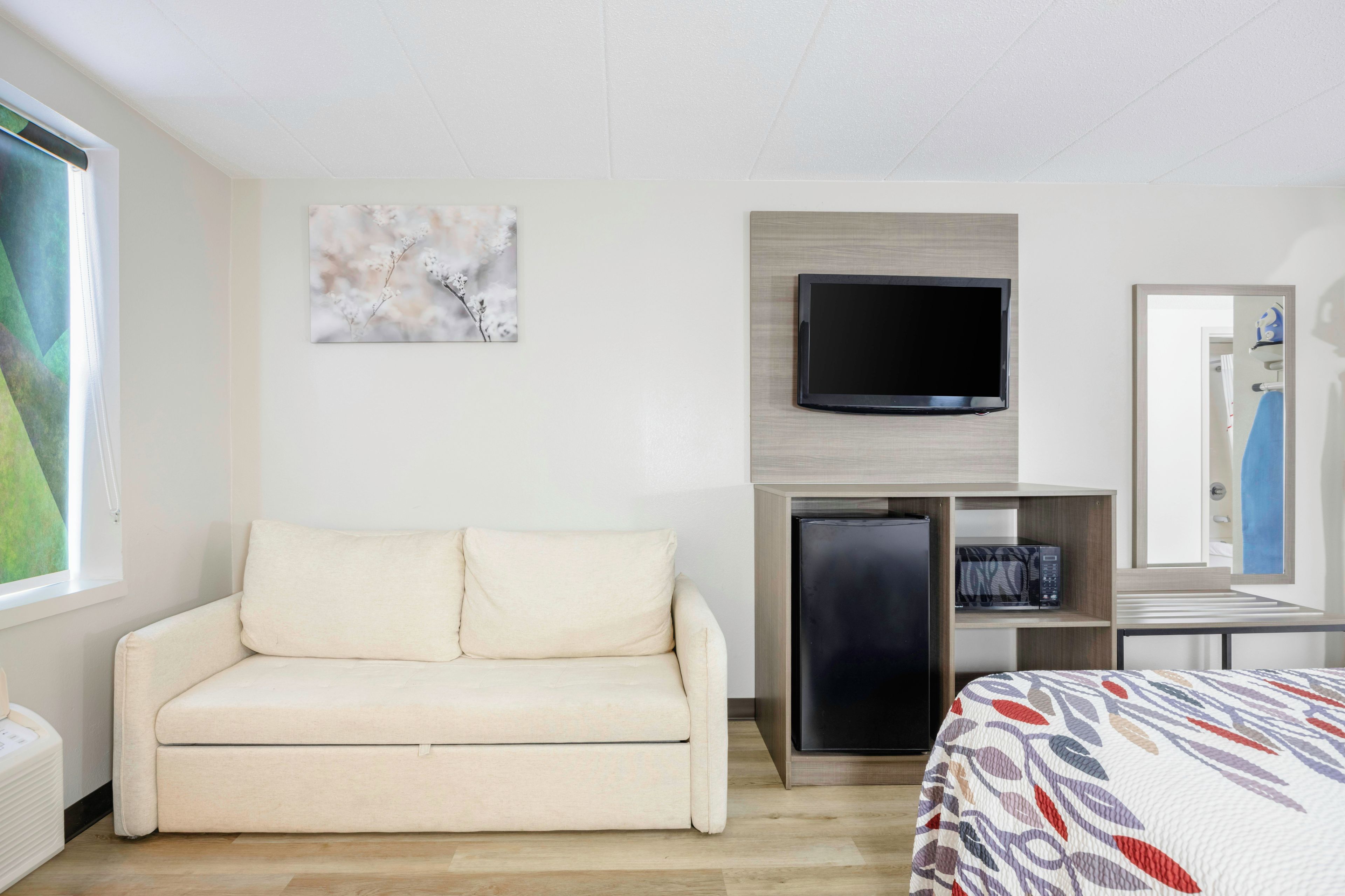 A hotel room with a sofa, a flat-screen TV mounted on the wall, a small refrigerator, and a bed with a patterned bedspread.