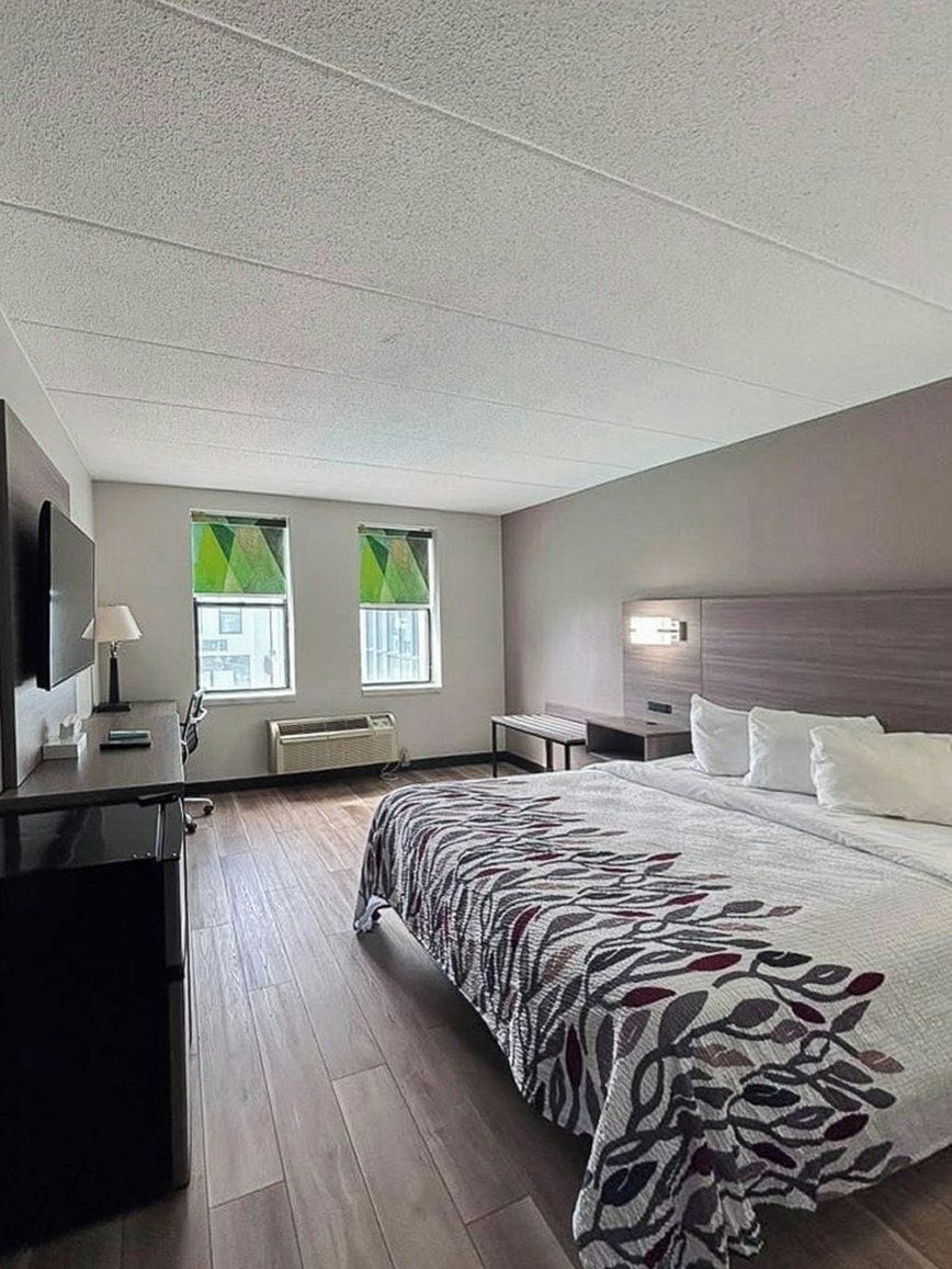 A hotel room with a large bed, a desk, a chair, a TV, and two windows with curtains.