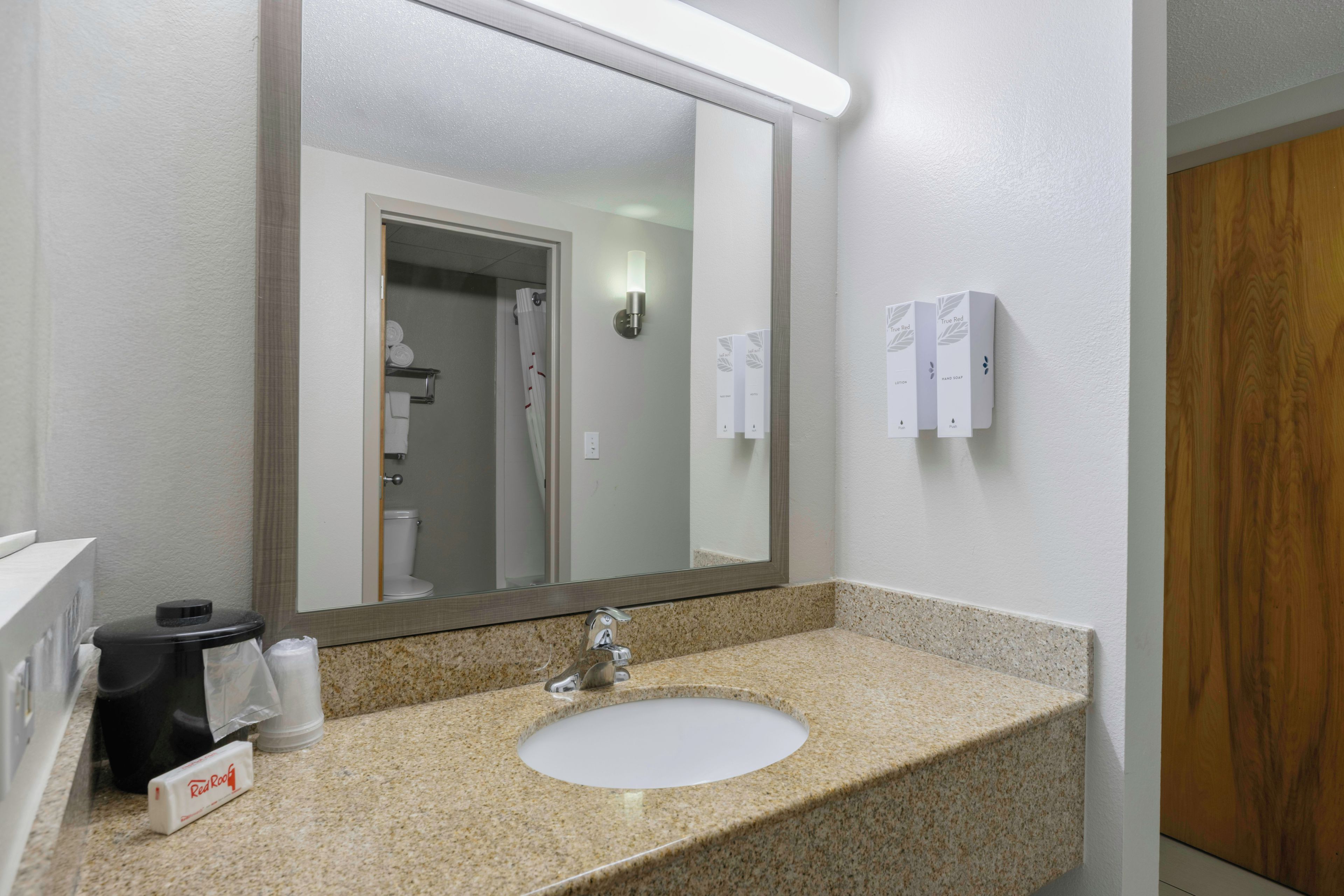 A bathroom with a granite countertop sink, a mirror, a towel dispenser, and a hand soap dispenser.