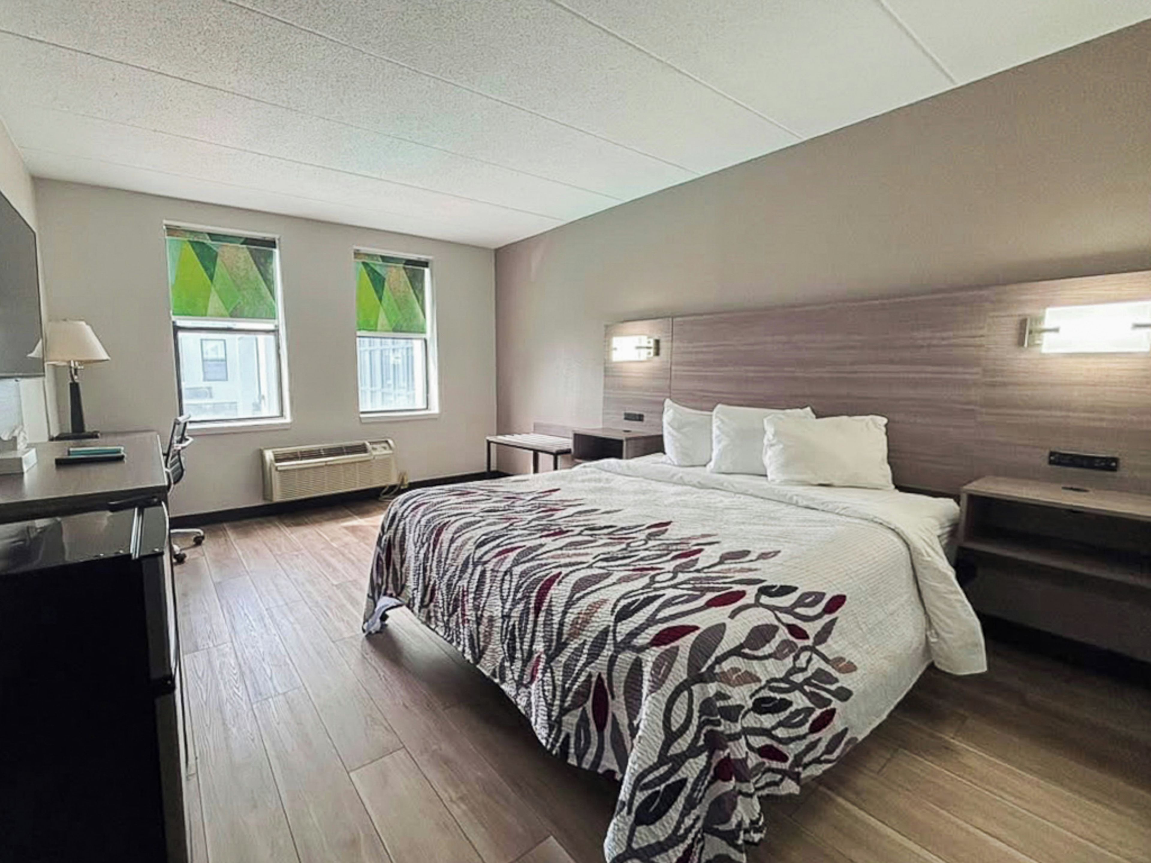 Una habitación de hotel con cama, mesitas de noche, escritorio y una silla. Hay dos ventanas con cortinas verdes.