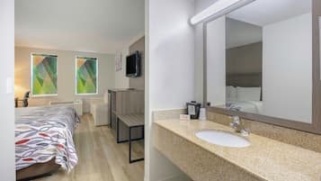 Habitación de hotel con cama, escritorio, silla, televisión, baño con lavamanos y espejo, y ventana con persianas.