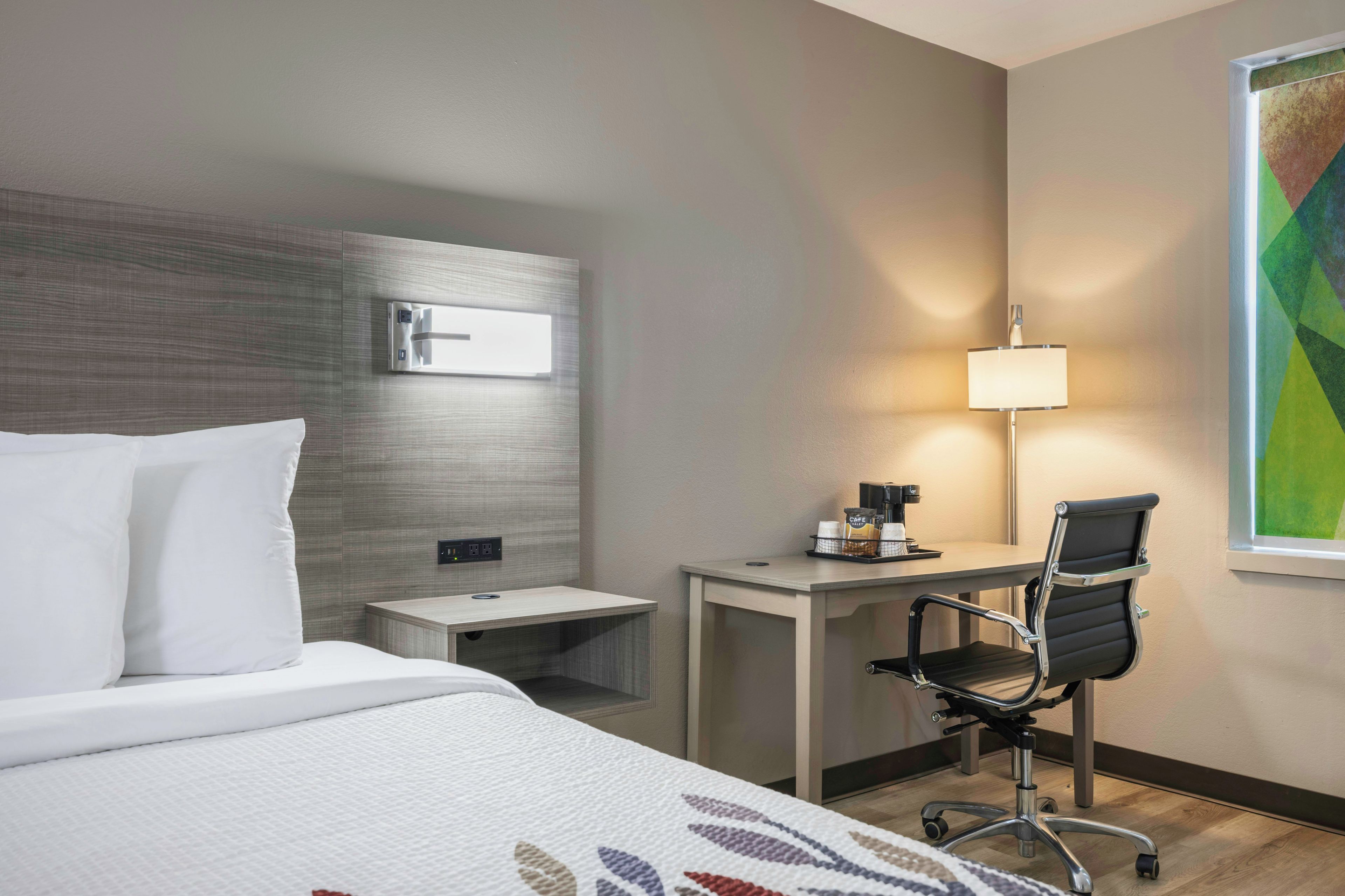 Ein Hotelzimmer mit Bett, Nachttisch, Schreibtisch, Stuhl, Lampe und einem Bild an der Wand.