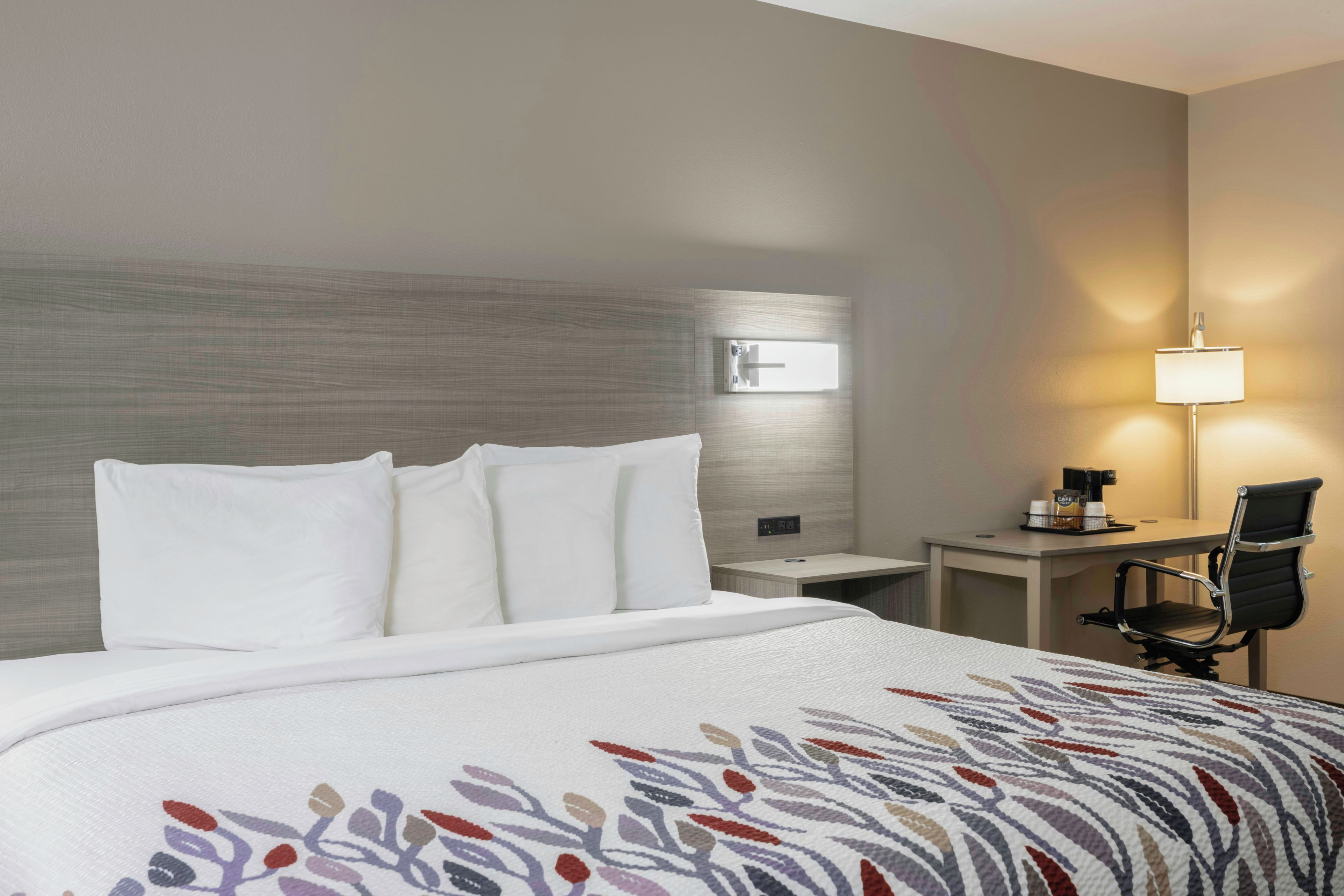 Habitación de hotel con cama, escritorio, silla, lámpara y toma de corriente en la pared.