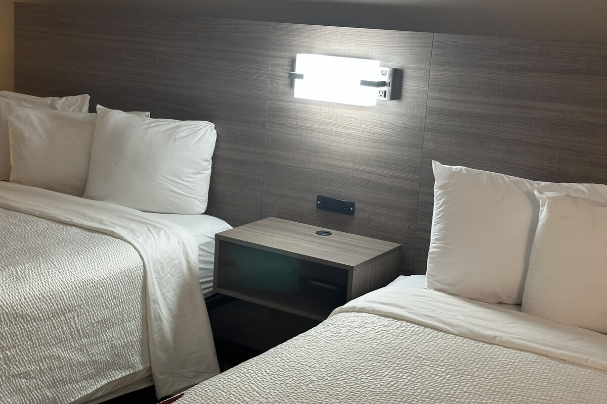 A hotel room with two beds, a nightstand, and a wall-mounted light.