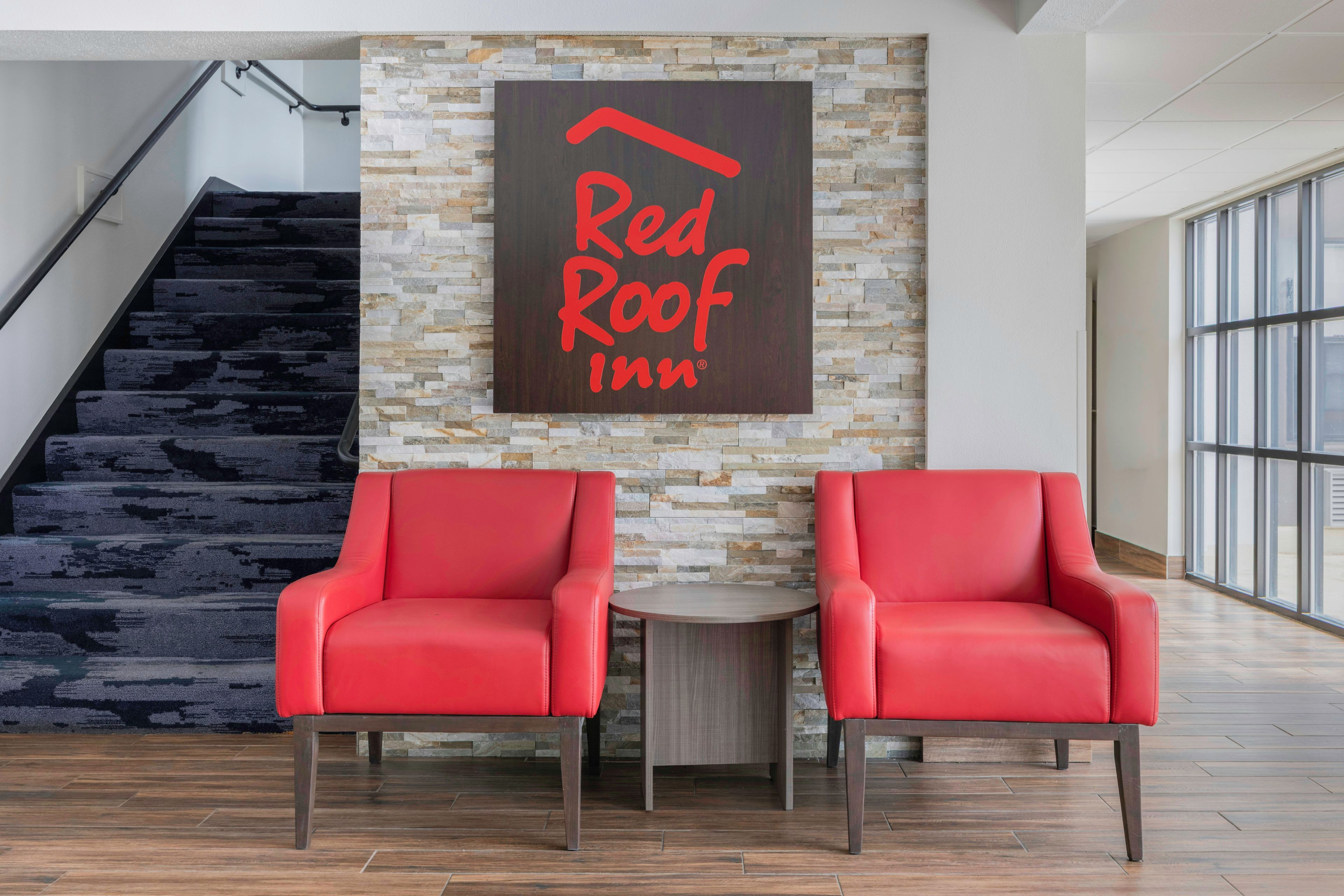 Un hall de hotel con una escalera, dos sillones de cuero rojo, una mesita y una pared con un letrero que dice 'Red Roof Inn'.