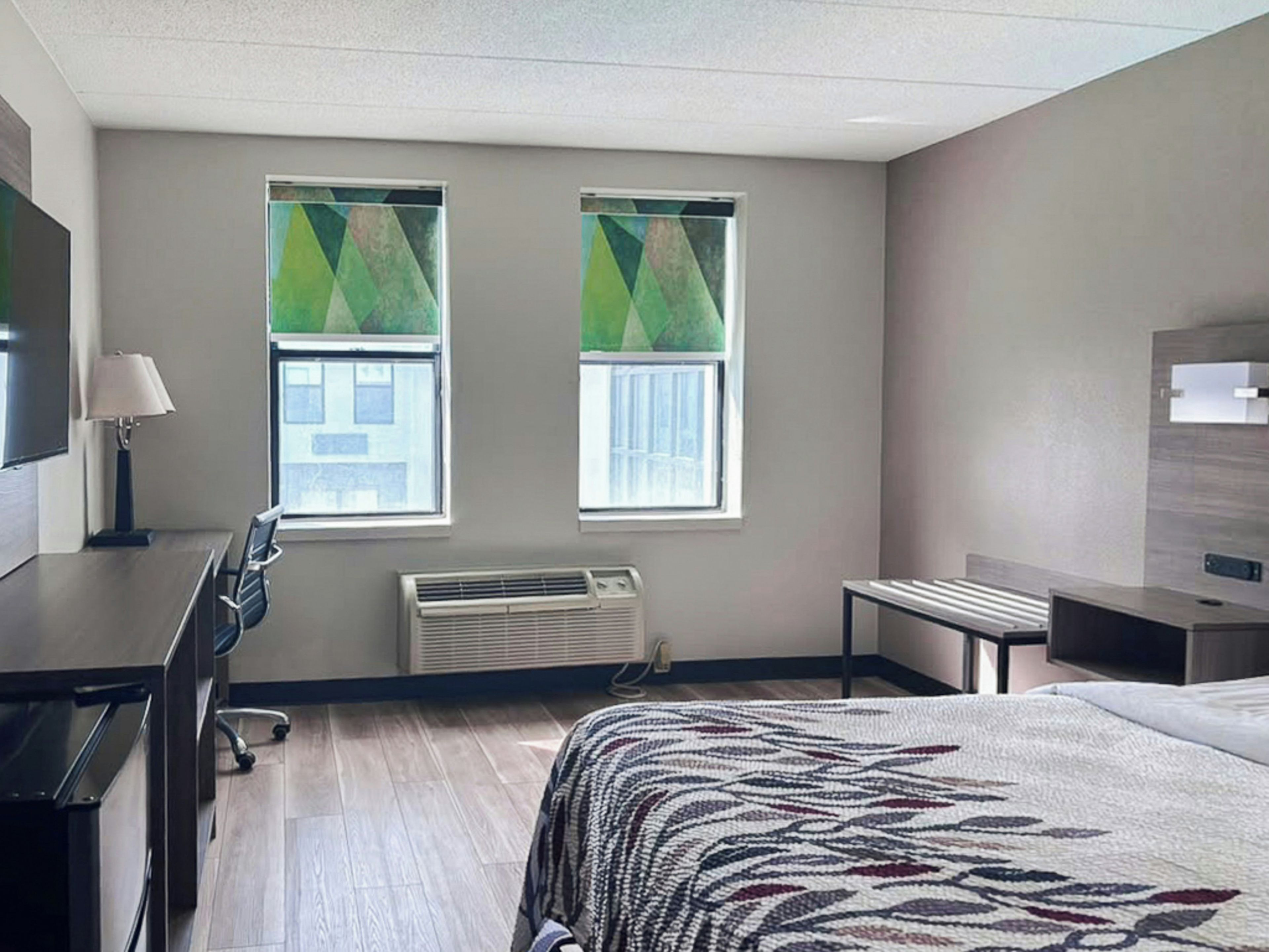 Habitación de hotel con cama, escritorio, silla, dos ventanas con persianas, unidad de aire acondicionado y televisor.