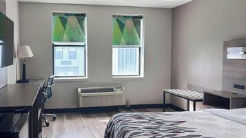 Habitación de hotel con cama, escritorio, silla, dos ventanas con persianas, unidad de aire acondicionado y televisor.