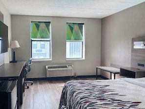 A hotel room with a bed, desk, chair, two windows with blinds, an air conditioning unit, and a television.