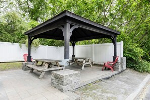 A covered outdoor seating area with picnic tables and benches, surrounded by a white fence and lush greenery.