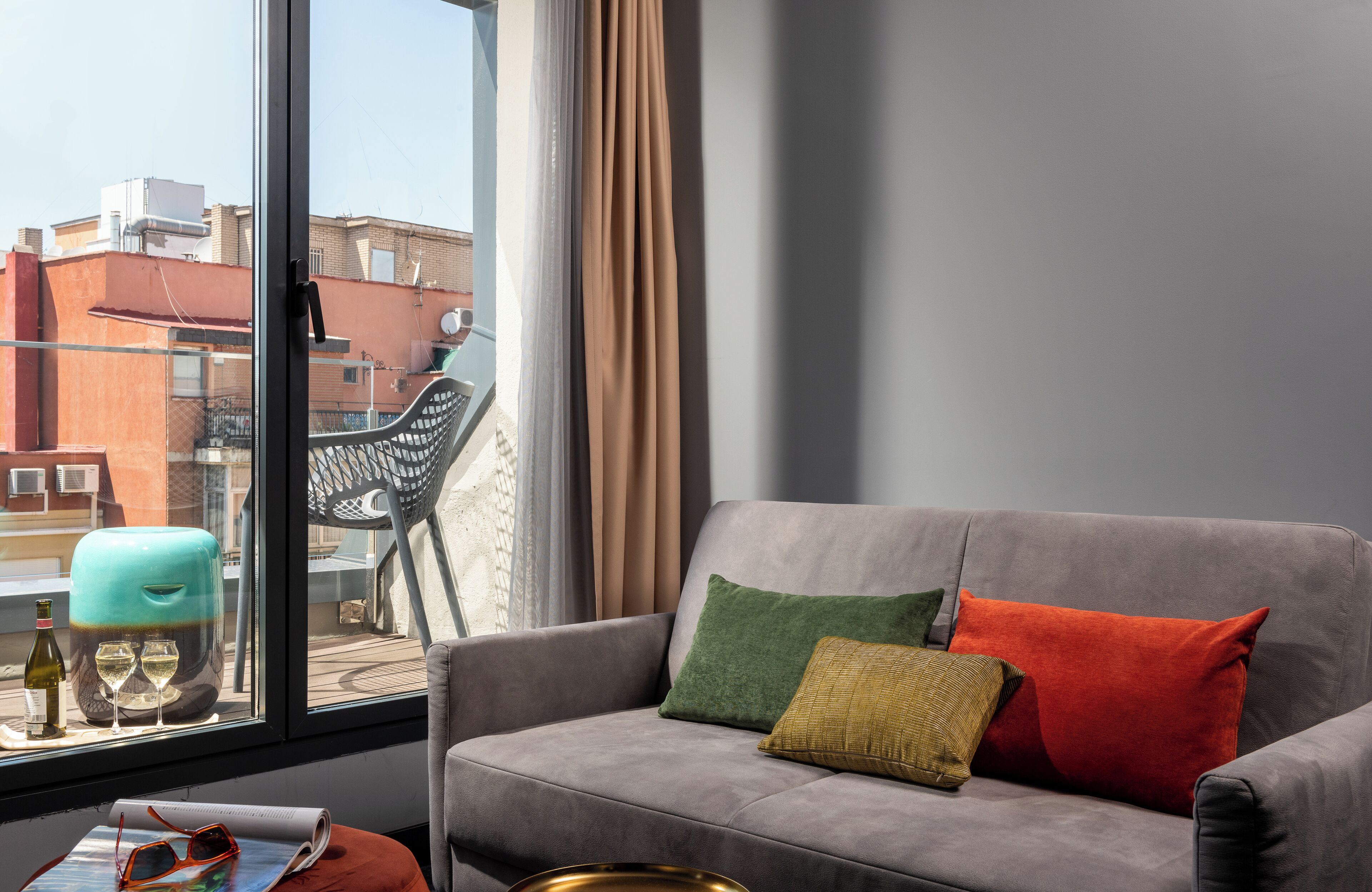 Ein modernes Wohnzimmer mit grauem Sofa, einem Fenster mit Blick auf Gebäude und einem Tisch mit Flasche und Gläsern.