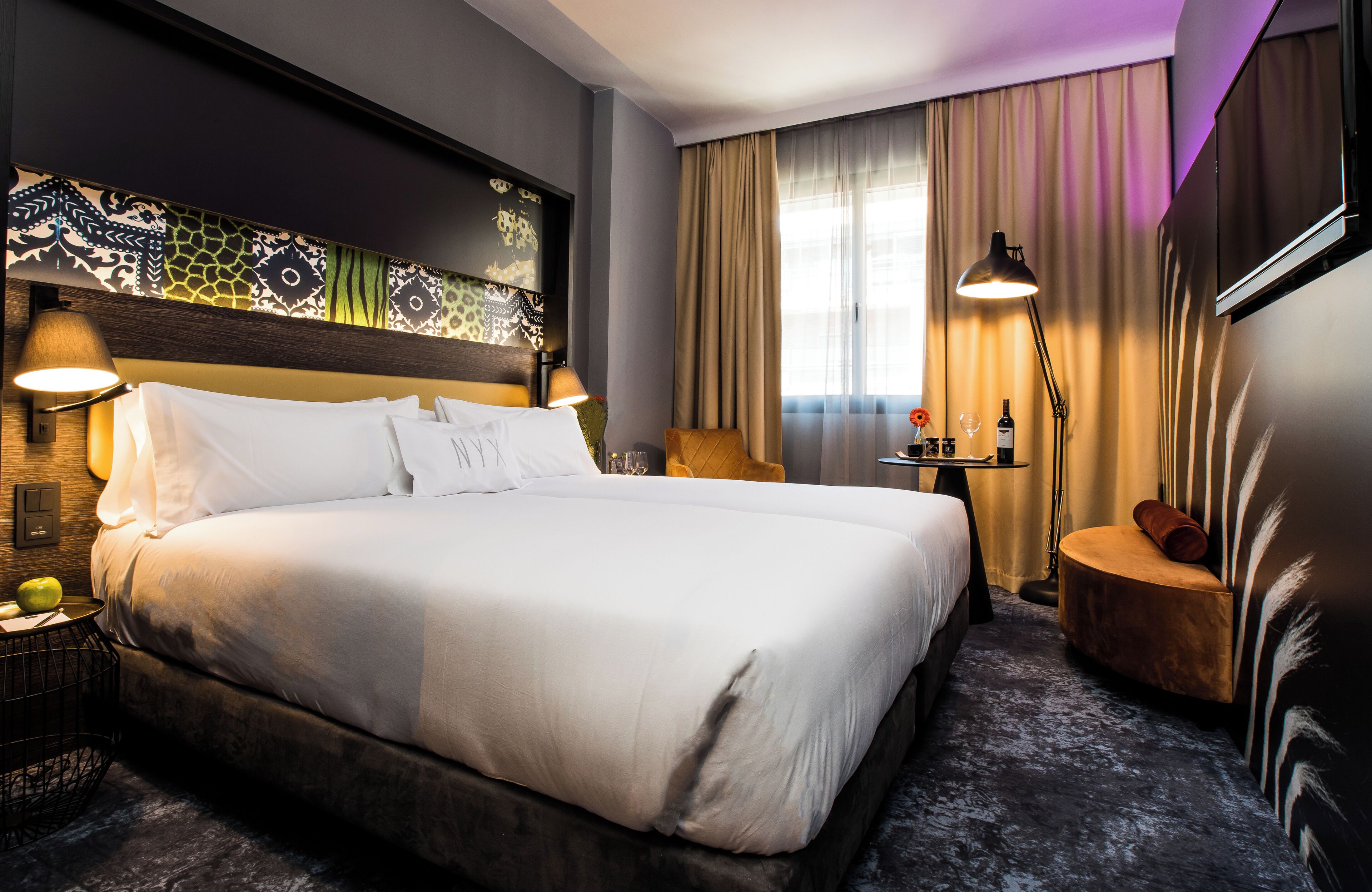Una habitación de hotel con una cama grande, cabecera con panel decorativo, mesita de noche con lámpara, una pequeña mesa redonda con lámpara y un ventanal con cortinas.