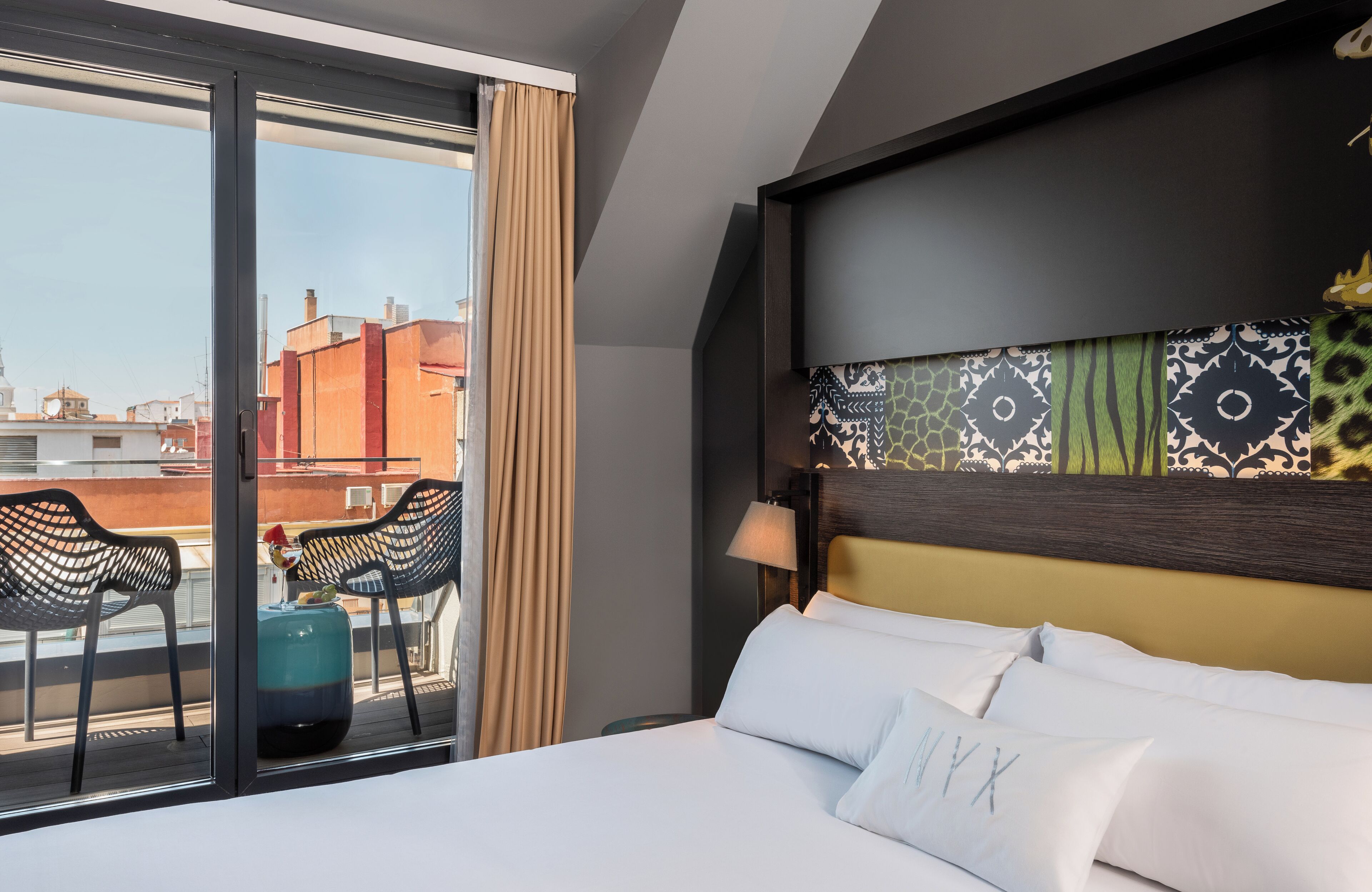 A hotel room with a bed, a window with curtains, a view of buildings, and a balcony with chairs and a table.