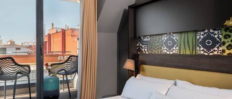 A hotel room with a bed, a window with curtains, a view of buildings, and a balcony with chairs and a table.