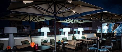 An outdoor seating area with tables and chairs under large umbrellas, with city views in the background.