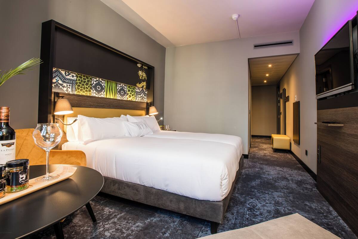 Camera d'albergo moderna con un letto grande, lampade ai lati del letto, un tavolino con bottiglia di vino e bicchiere, e una testiera decorativa.