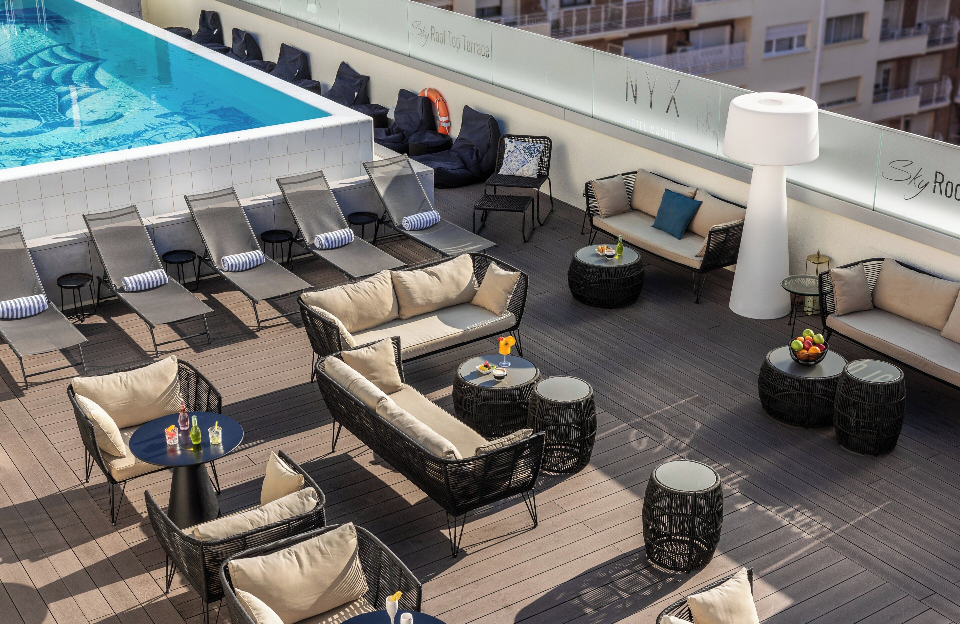 A rooftop lounge area with sun loungers, sofas, and round tables, overlooking a swimming pool.