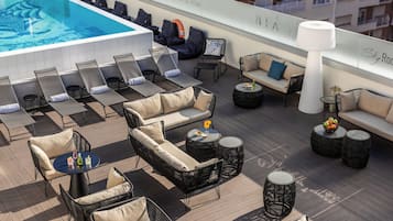A rooftop lounge area with sun loungers, sofas, and round tables, overlooking a swimming pool.