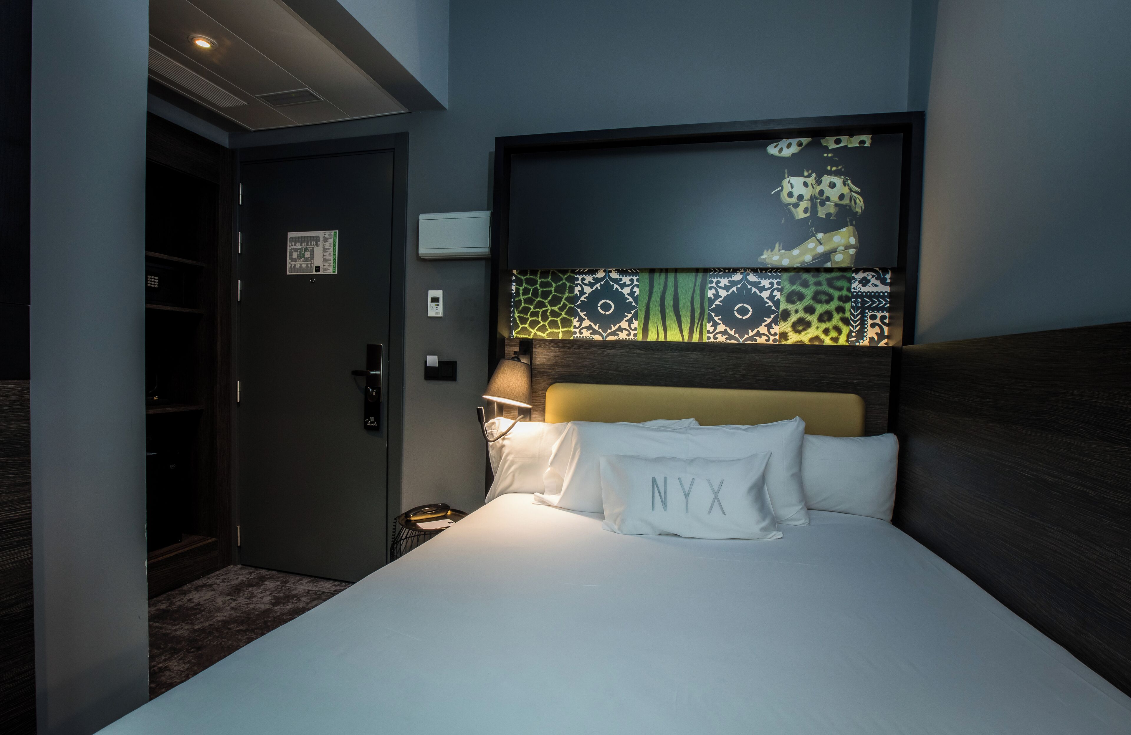 Habitación de hotel con cama, cabecera con panel decorativo, mesita de noche con lámpara y puerta que da a un armario.