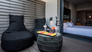 A modern hotel room with a bed, a seating area with black cushions, a small round table with a fruit tray, and a minibar.