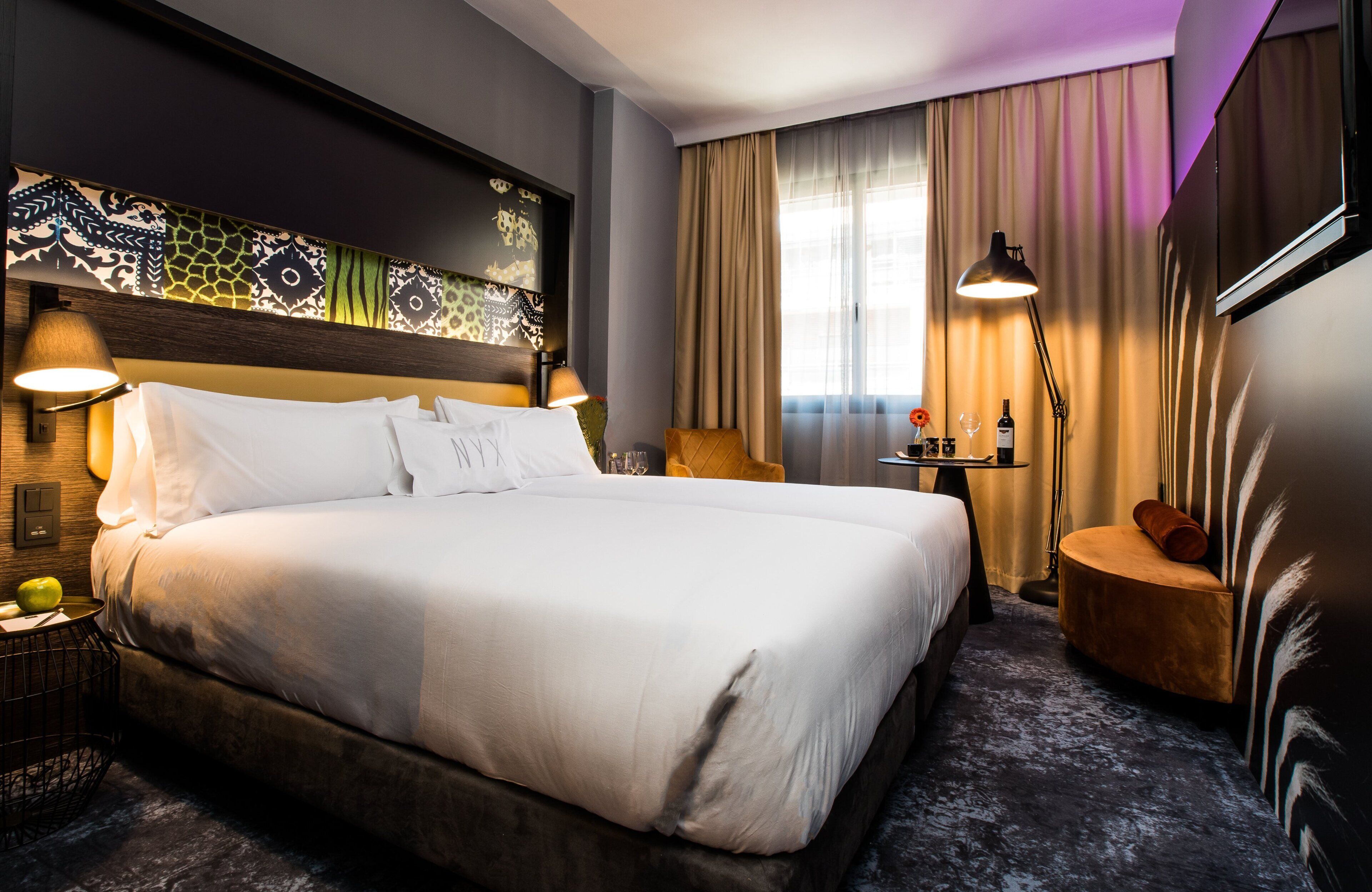 A hotel room with a large bed, a nightstand, a lamp, and a view of the city through the window.