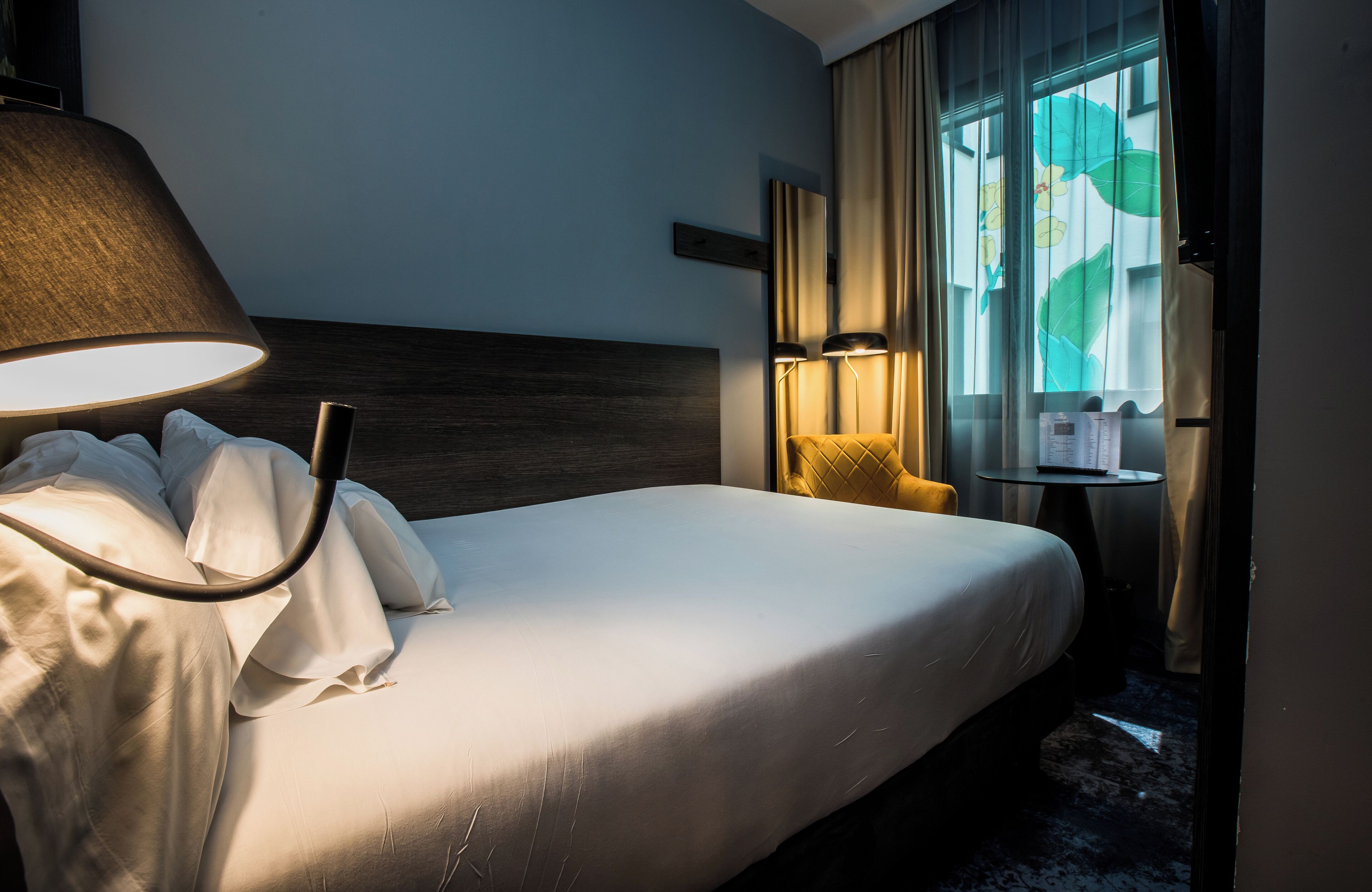 Ein Hotelzimmer mit einem großen Bett, einer Nachttischlampe, einem kleinen Tisch inklusive Lampe und einem Fenster mit Vorhängen.