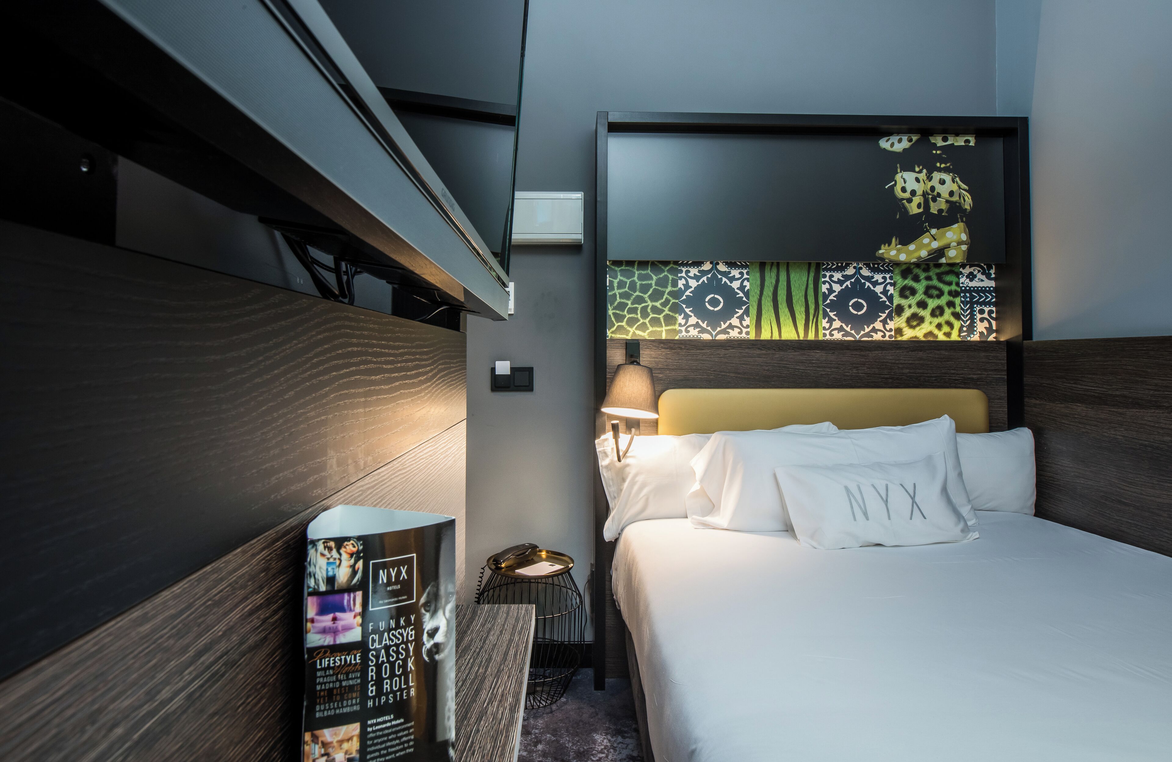 A modern hotel room with a large bed, a headboard with patterned designs, a bedside table with a lamp, and a framed artwork above the bed.