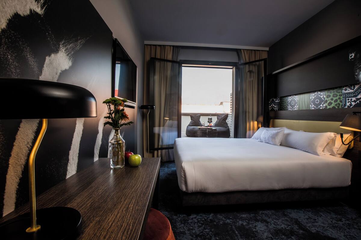 Una moderna camera d'hotel con un ampio letto, una scrivania con lampada, un vaso con fiori e vista sulla città dalla finestra.