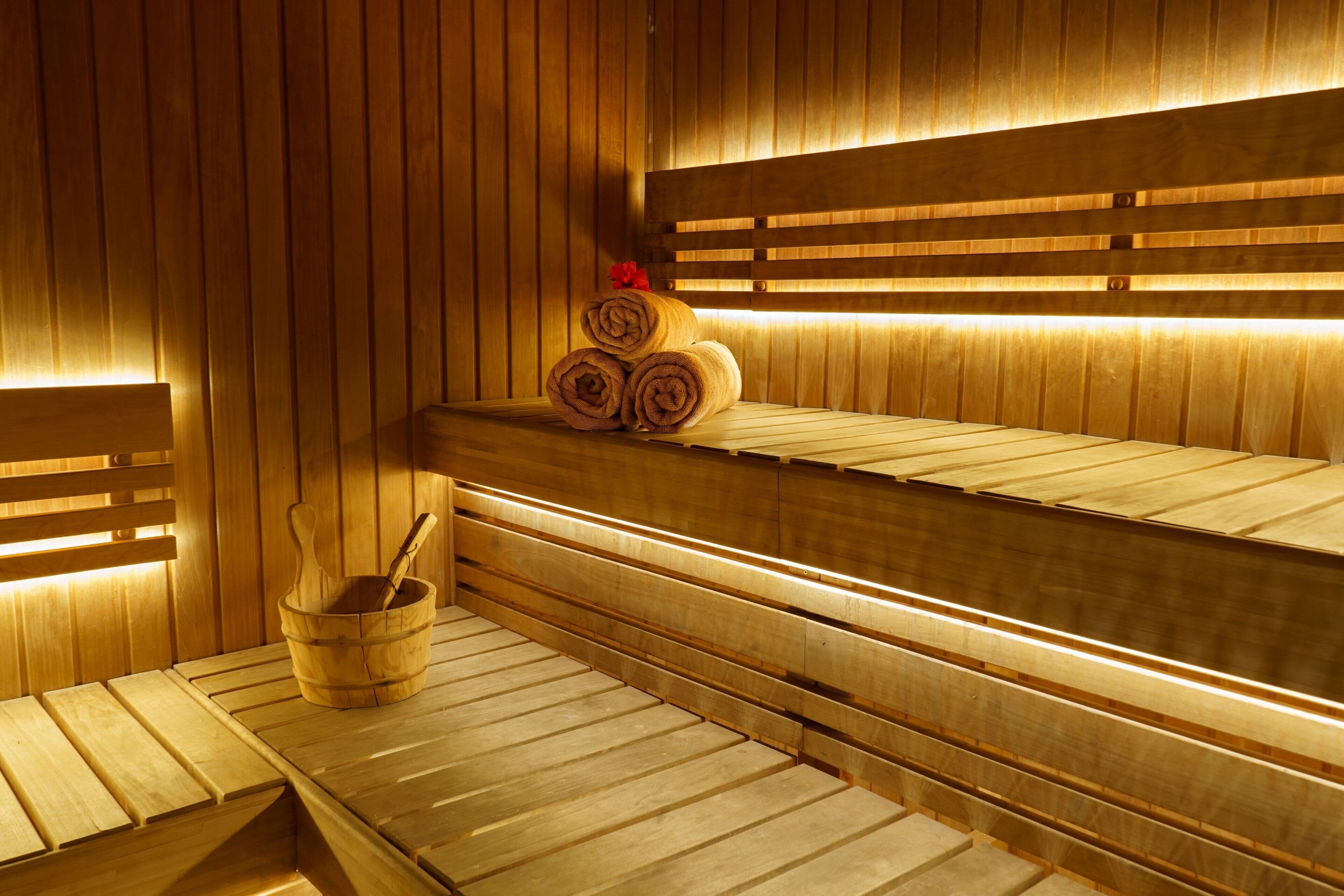A sauna with wooden benches and benches, a wooden bucket with a brush, and rolled towels on a bench.