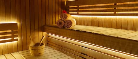 A sauna with wooden benches and benches, a wooden bucket with a brush, and rolled towels on a bench.