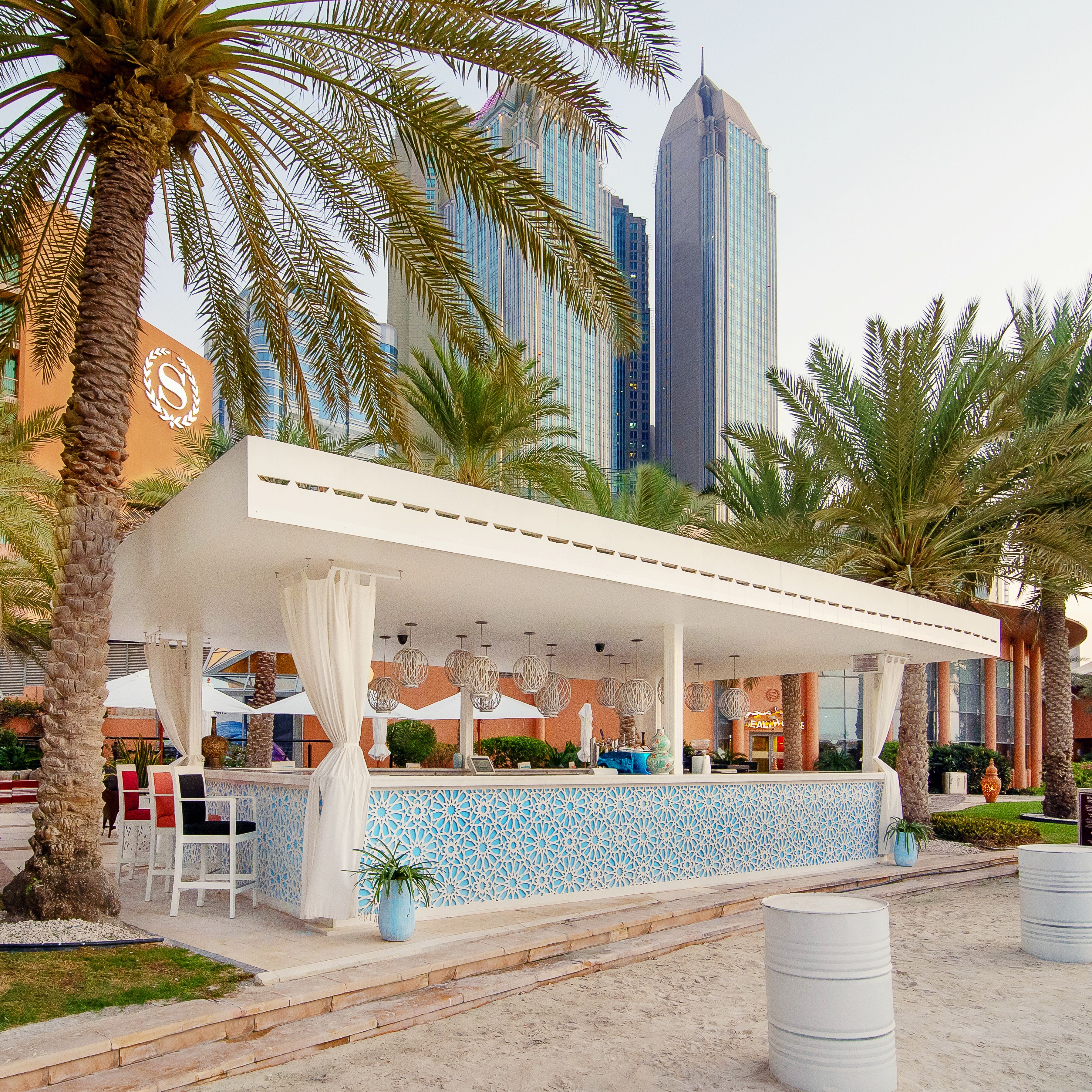 A covered outdoor bar area with a white canopy, palm trees, and a city skyline in the background.