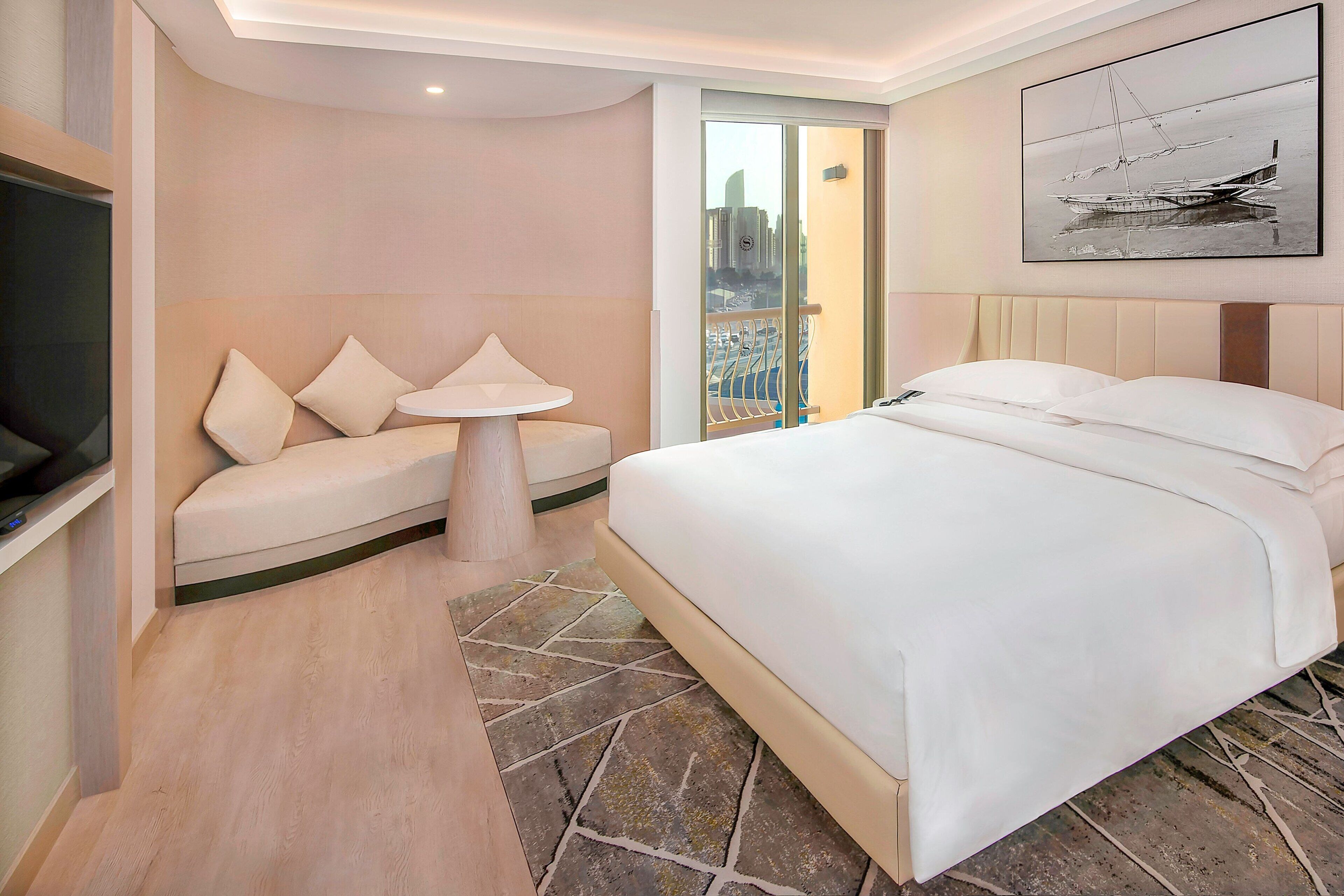 A hotel room with a bed, a sofa, a small table, and a view of the city through a glass door.