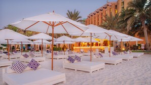 A beachside lounge area with white sunbeds and umbrellas, a bar in the background, and palm trees. - Sheraton Abu Dhabi Hotel & Resort (Abu Dhabi)