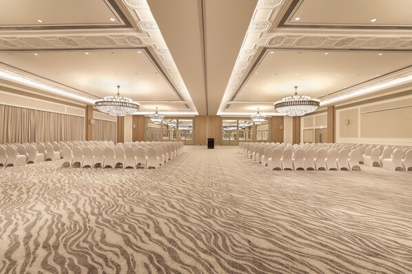 Un amplio salón de conferencias con filas de sillas blancas, alfombra estampada y candelabros ornamentados.