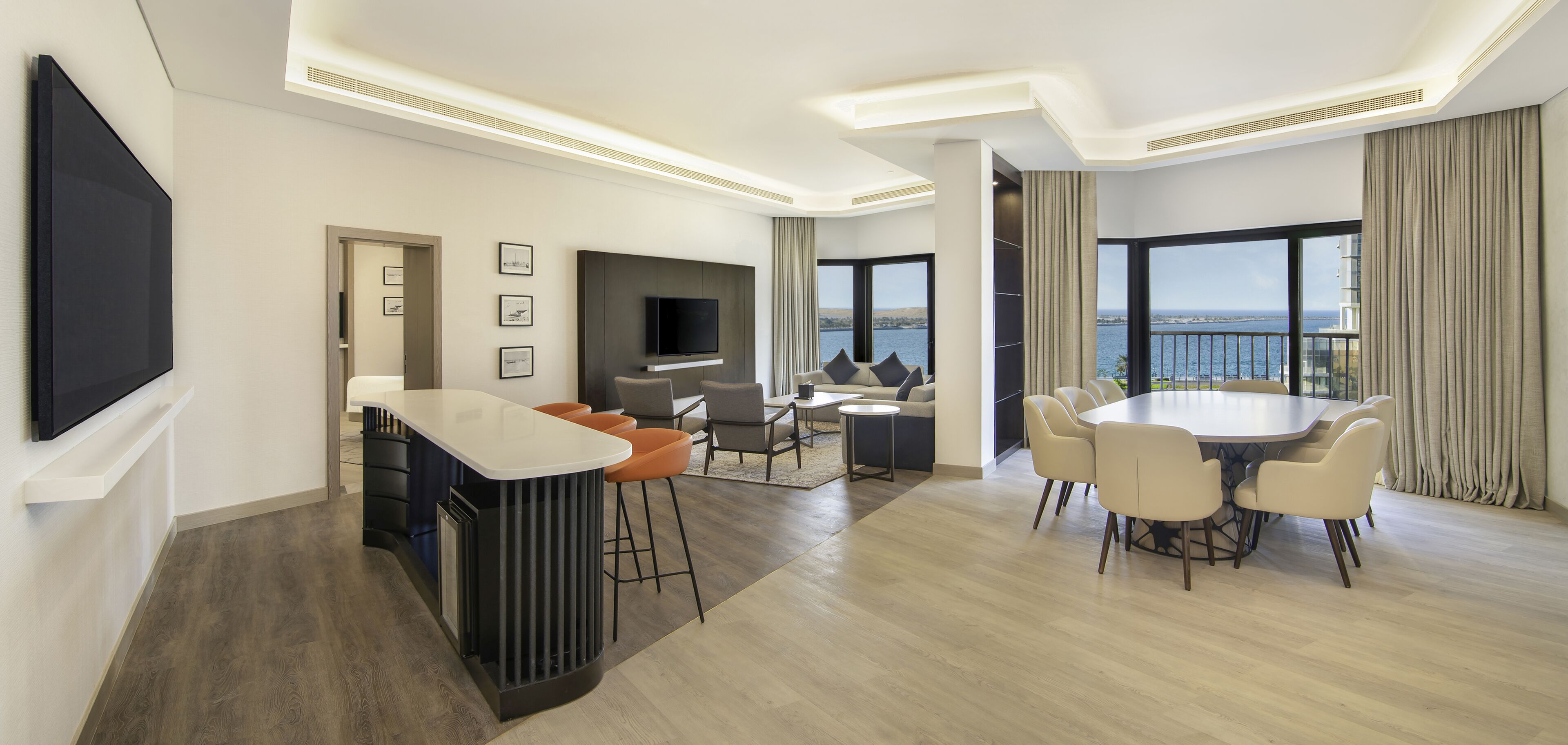 A modern hotel room with a dining area, a bar, and a large window offering a view of the sea.