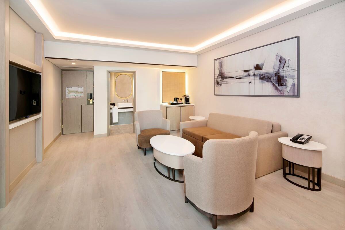 Una habitación de hotel moderna con un sofá, sillones, una mesa de centro, una mesita auxiliar con un teléfono, un televisor y un cuadro en la pared.