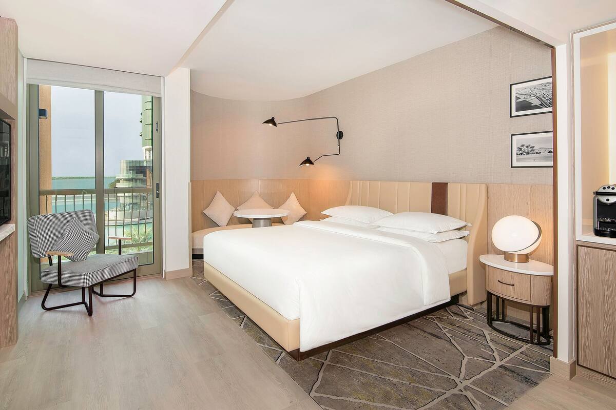Una habitación de hotel moderna con una cama grande, una zona de estar con una silla, una mesita y vistas al exterior.