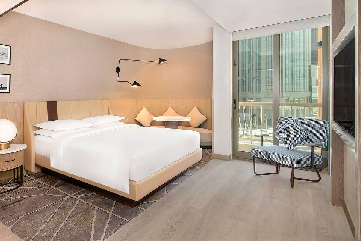 Una habitación de hotel moderna con una cama grande, una mesita, una silla y un balcón con vistas a la ciudad.