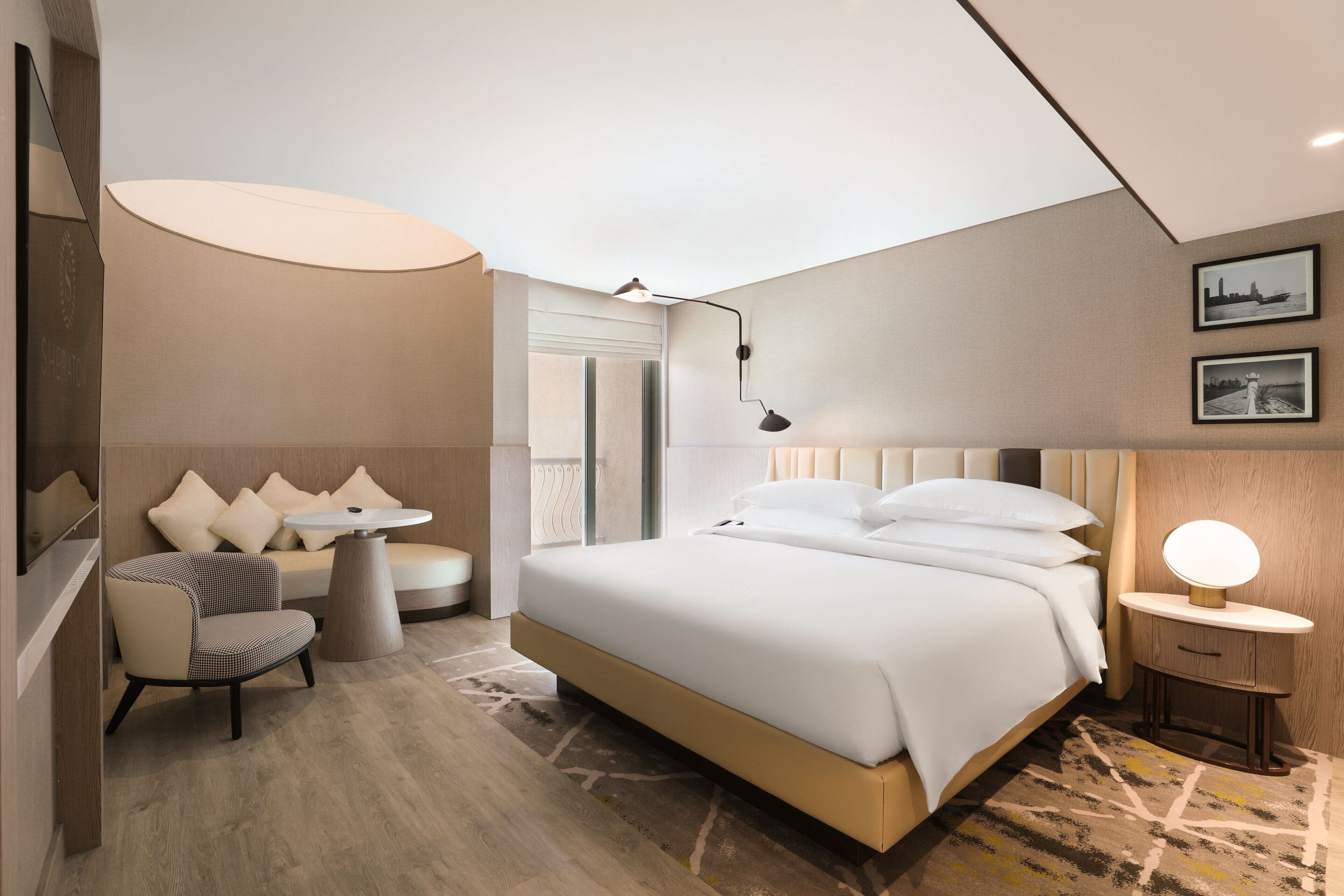 Ein modernes Hotelzimmer mit einem großen Bett, einer Sitzecke mit Tisch und einem kleinen Nachttisch mit Lampe.