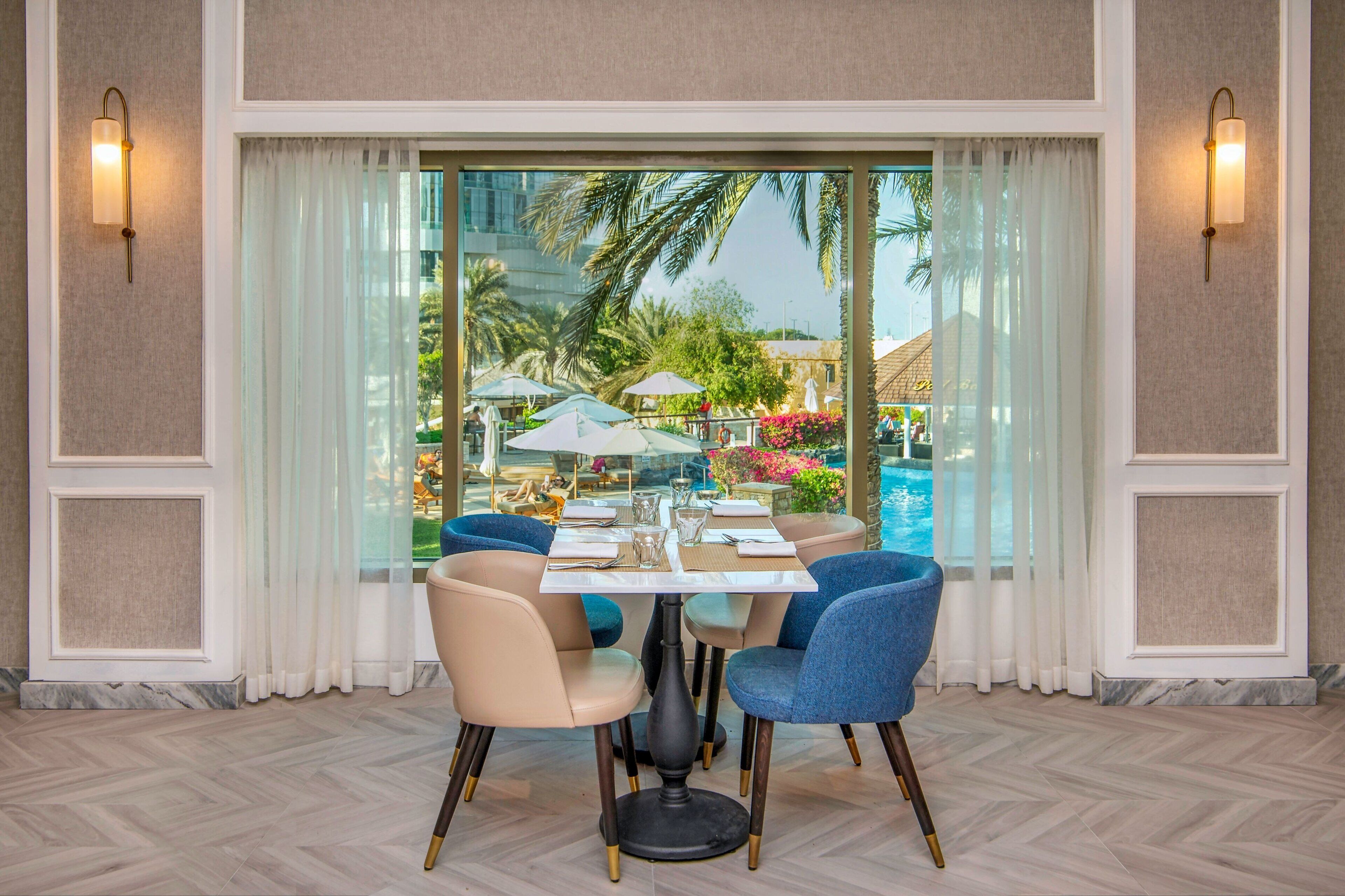 Un comedor con una mesa dispuesta para cuatro personas, ventanas amplias con cortinas y vistas a una piscina y palmeras.