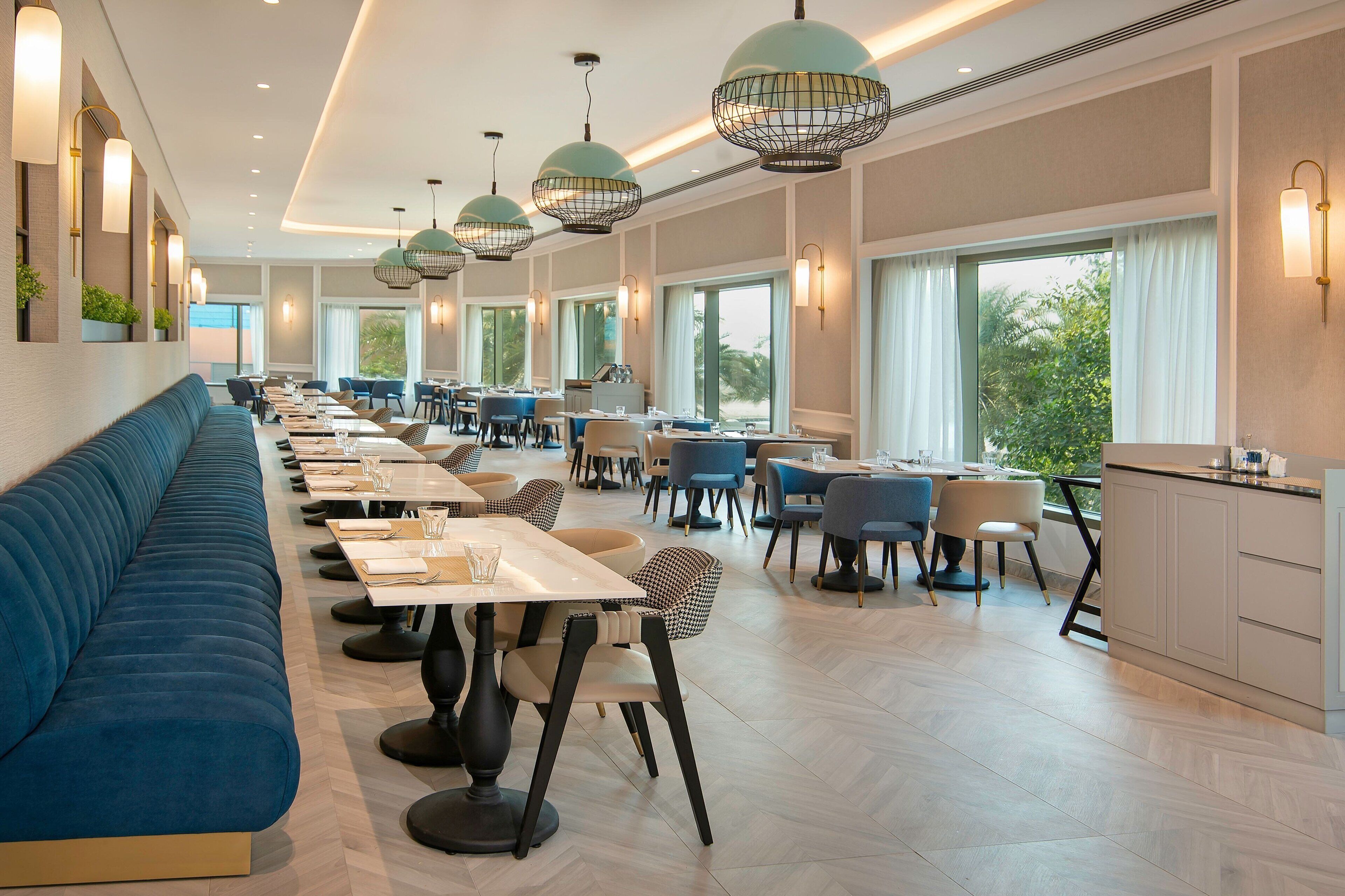 A modern restaurant with a long blue sofa, multiple dining tables, and pendant lighting.