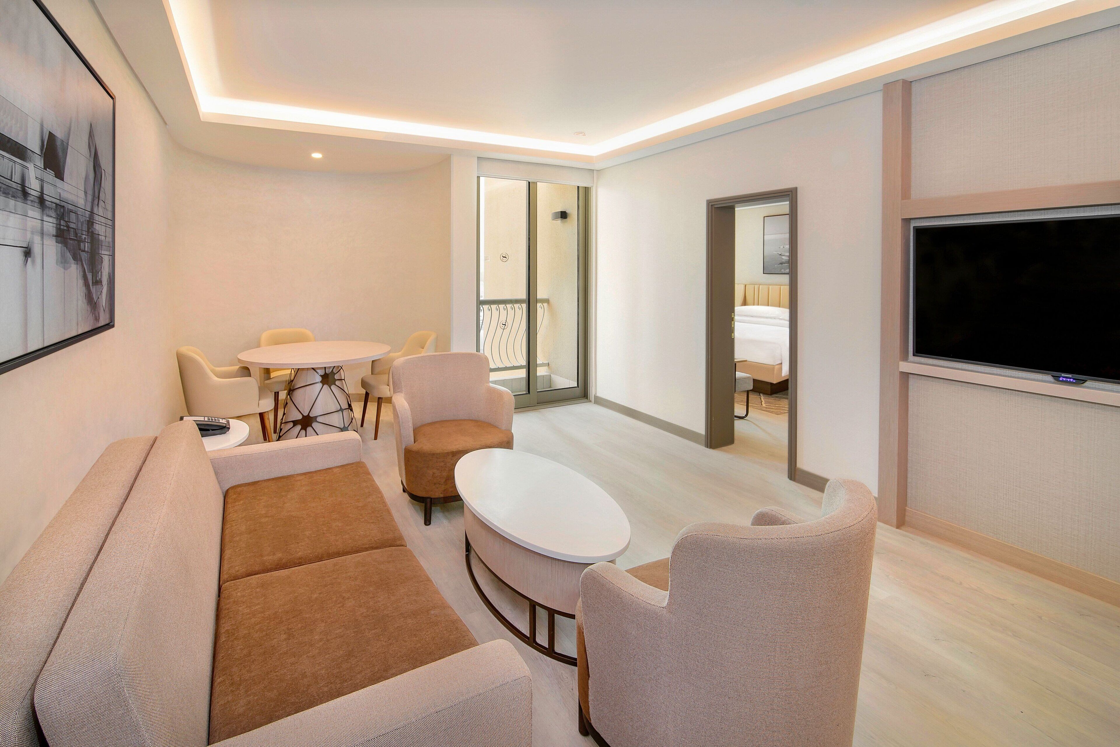 A modern hotel room with a sofa, two armchairs, a small round table, and a TV.