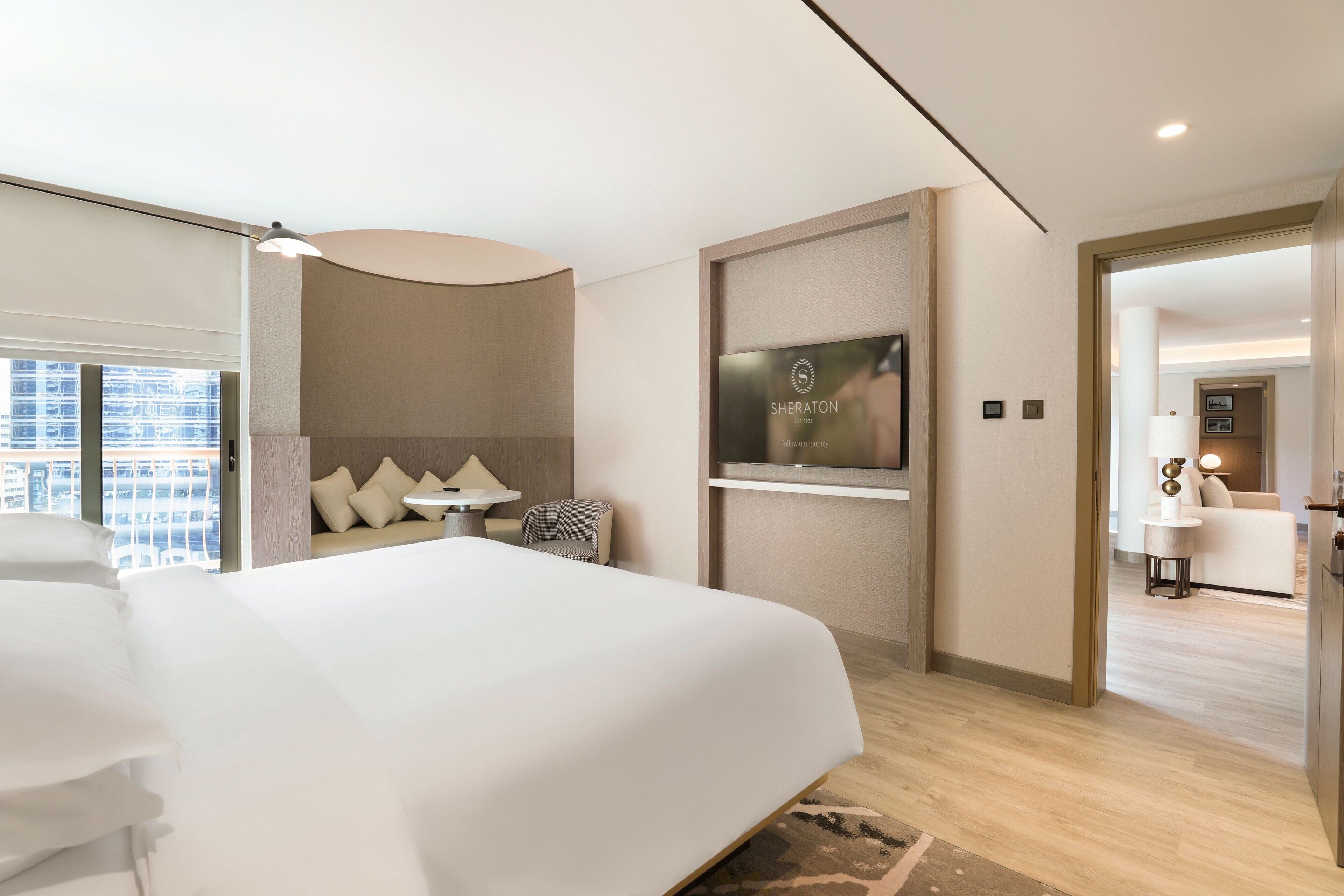 Ett modernt hotellrum med en stor säng, en sittgrupp med soffa och fåtöljer, en platt-TV som är monterad på väggen och utsikt över staden genom stora fönster.